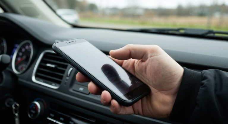 A close-up photo of a delivery driver's hand holding a smartphone inside a car, with soft morning light.