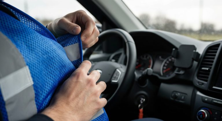 Close-up of a person wearing a blue and grey high-visibility safety vest with reflective stripes inside a car.