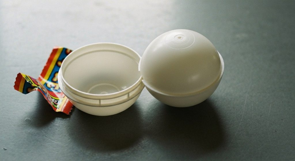 A disassembled white plastic toy capsule and its torn outer wrapper on a grey work surface.