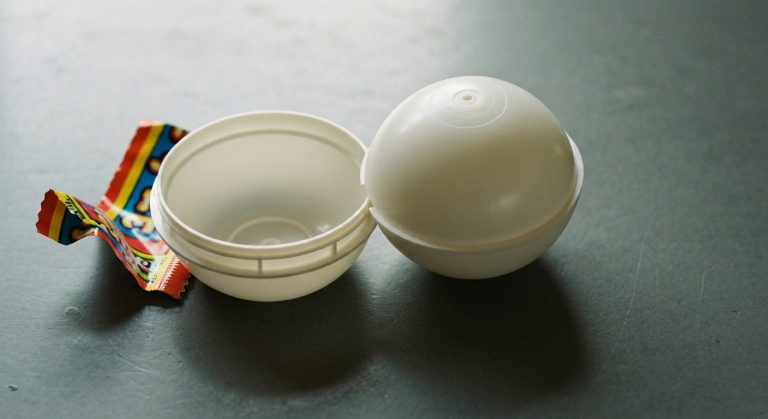A disassembled white plastic toy capsule and its torn outer wrapper on a grey work surface.