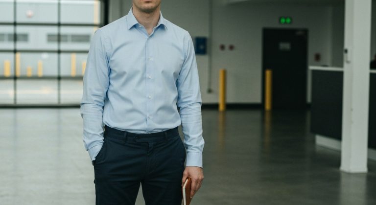 Warehouse Interview Attire: Ditch the Suit (2026) 14 A man in a light blue button-down shirt and dark chinos stands in a modern warehouse lobby holding a notebook.