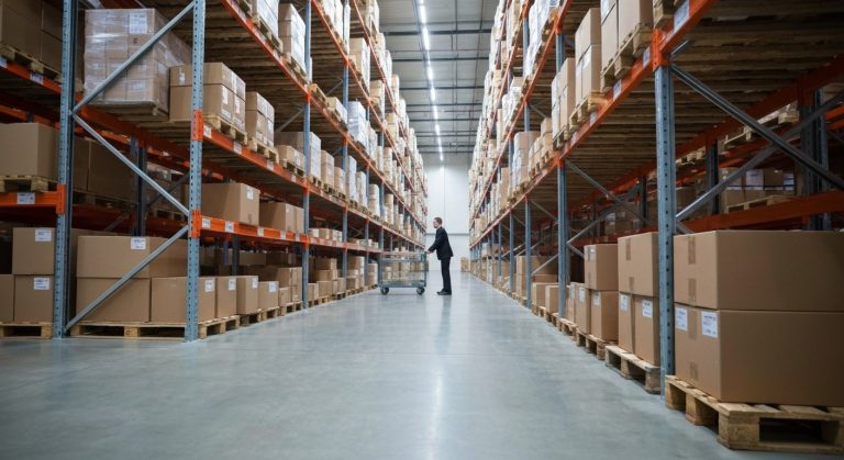 How to Be a Faster Warehouse Picker (No Running) 2 Wide shot of an organized warehouse aisle with a worker pushing a picking cart on a polished concrete floor.